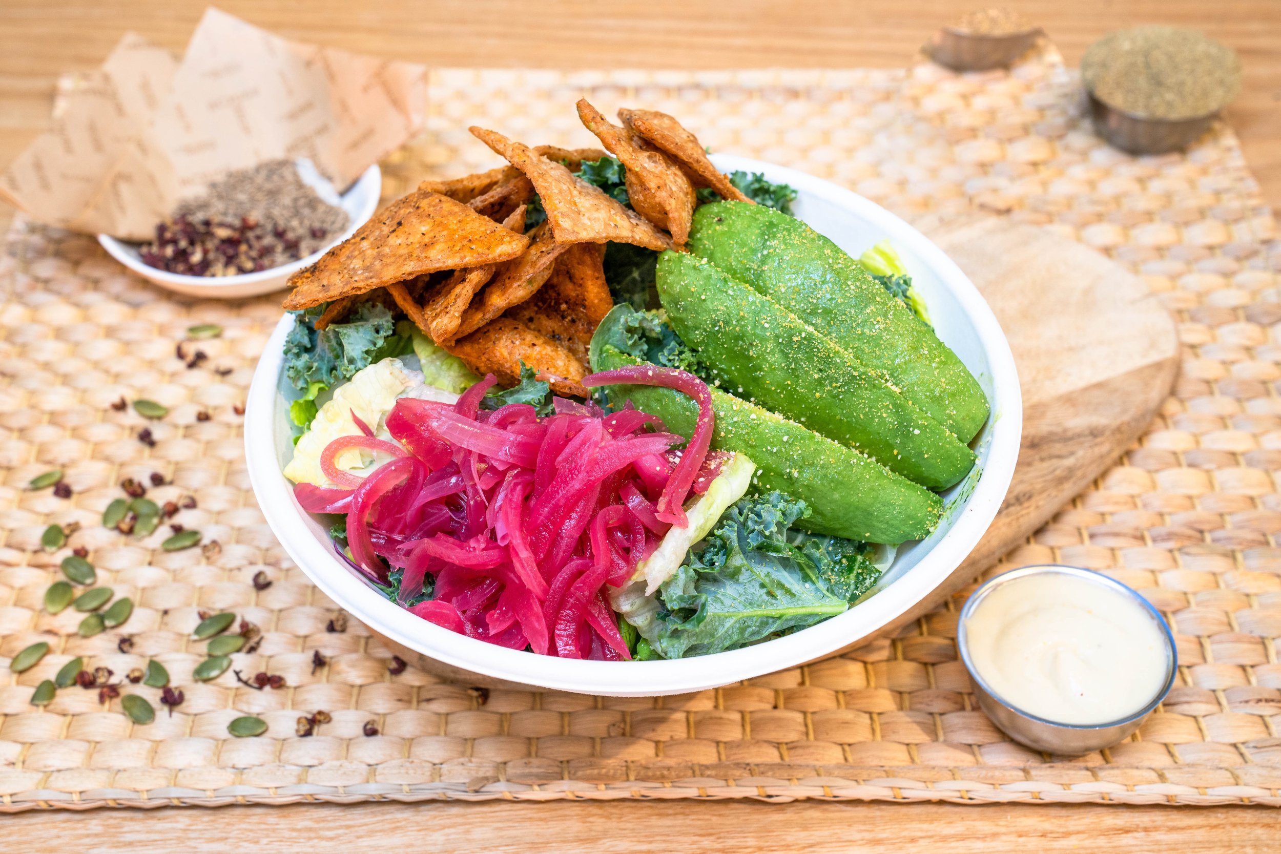 A salad bowl with avocado slices, kale, pickled onions, and triangular tortilla chips, served with a small cup of dressing, on a woven placemat with seeds scattered around.