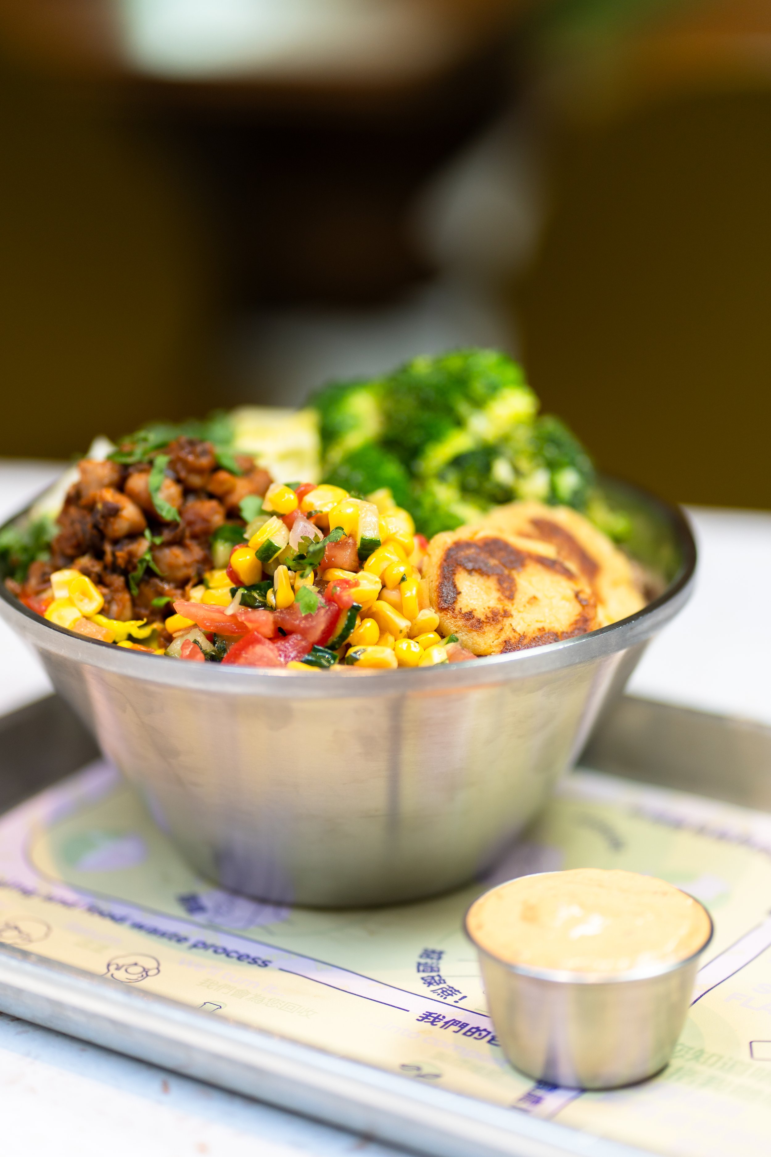 Vegetarian Buddha bowl with corn, tomatoes, black beans, broccoli, and naan bread, served with dipping sauce in a metal bowl on a tray.
