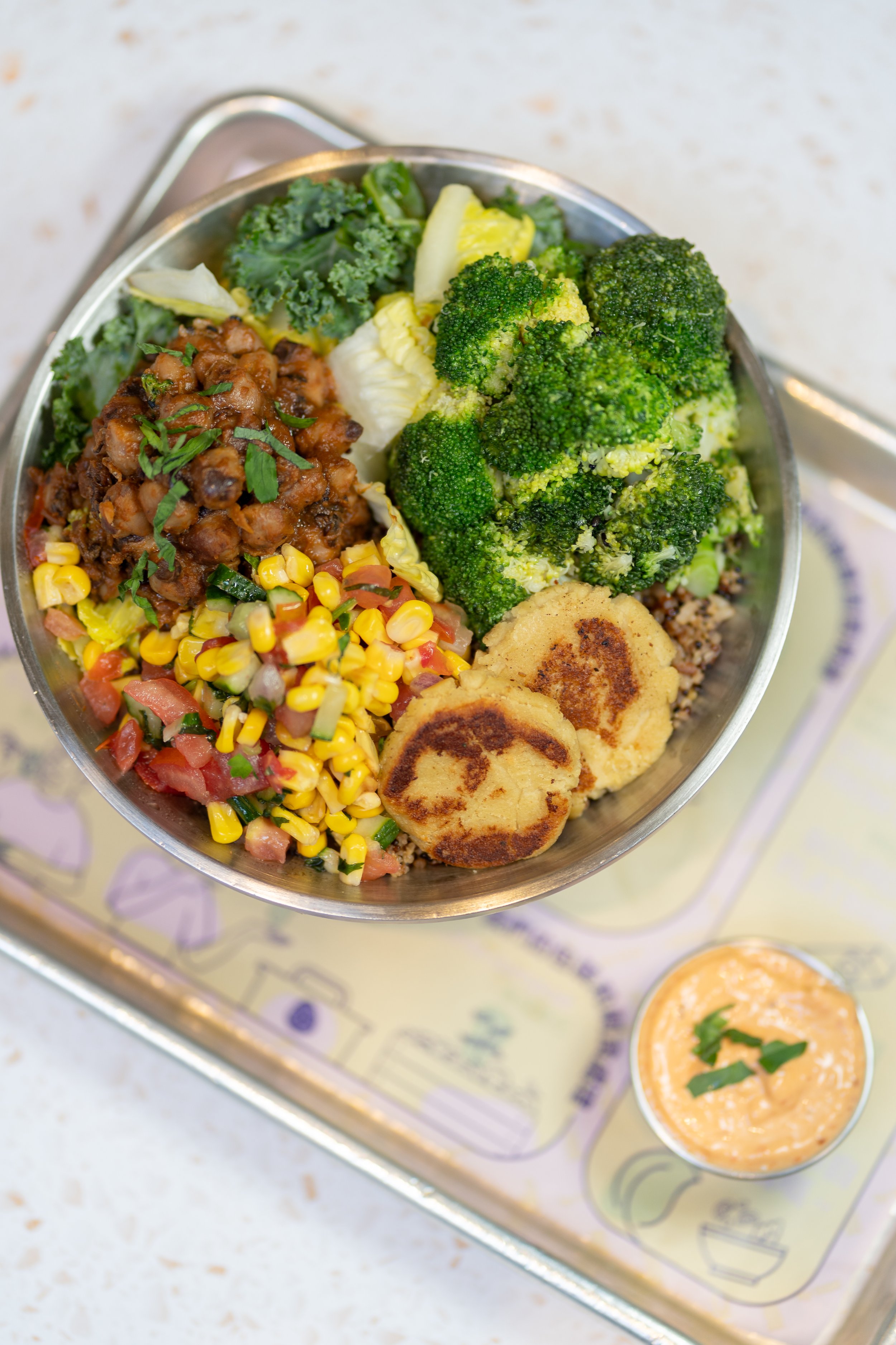 A bowl containing broccoli, kale, corn salad, black-eyed peas, and two patties, served with a side of creamy dipping sauce.