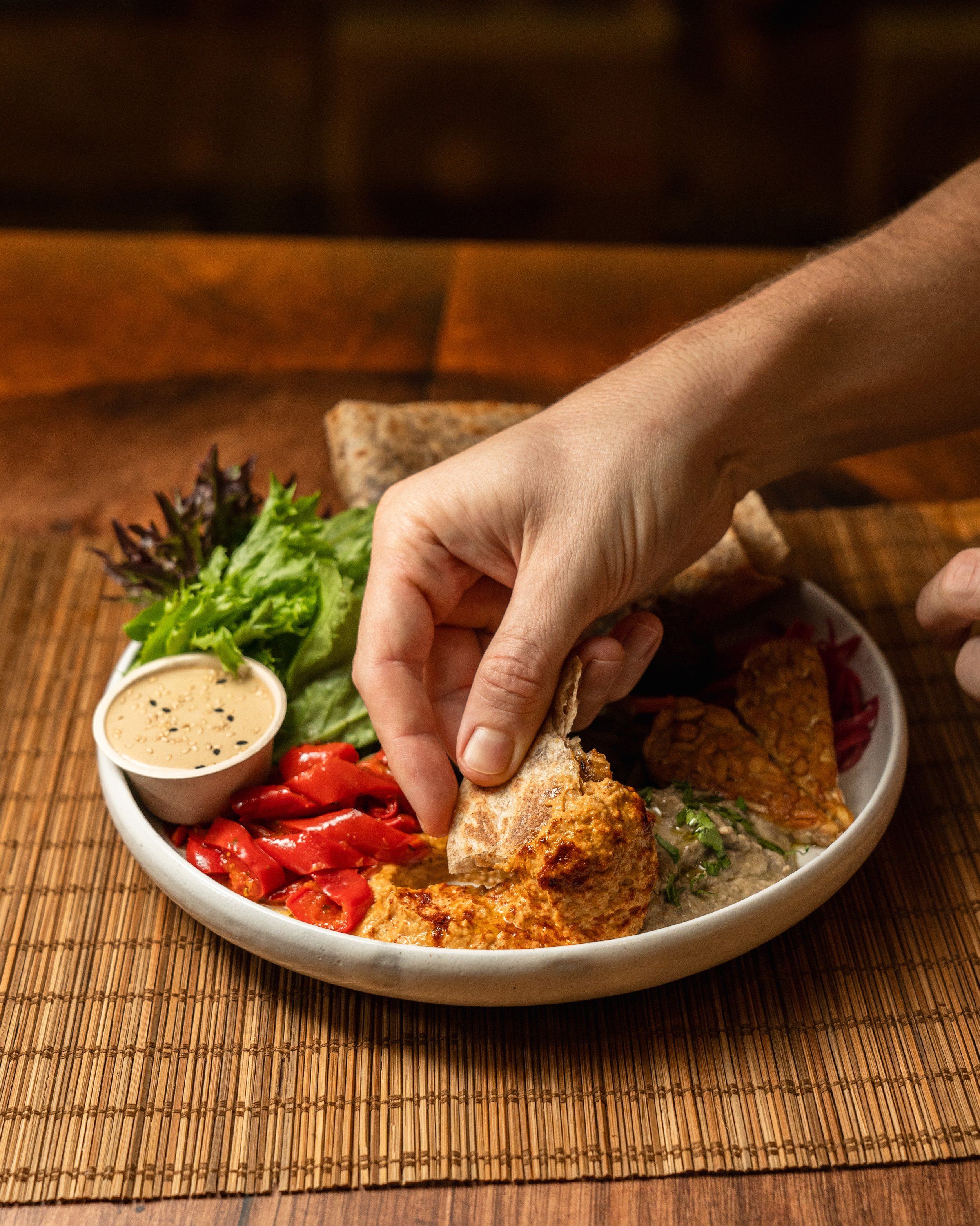 A person dipping bread into a plate with hummus, roasted red peppers, mixed greens, tempeh, and a small cup of dipping sauce on a bamboo mat.