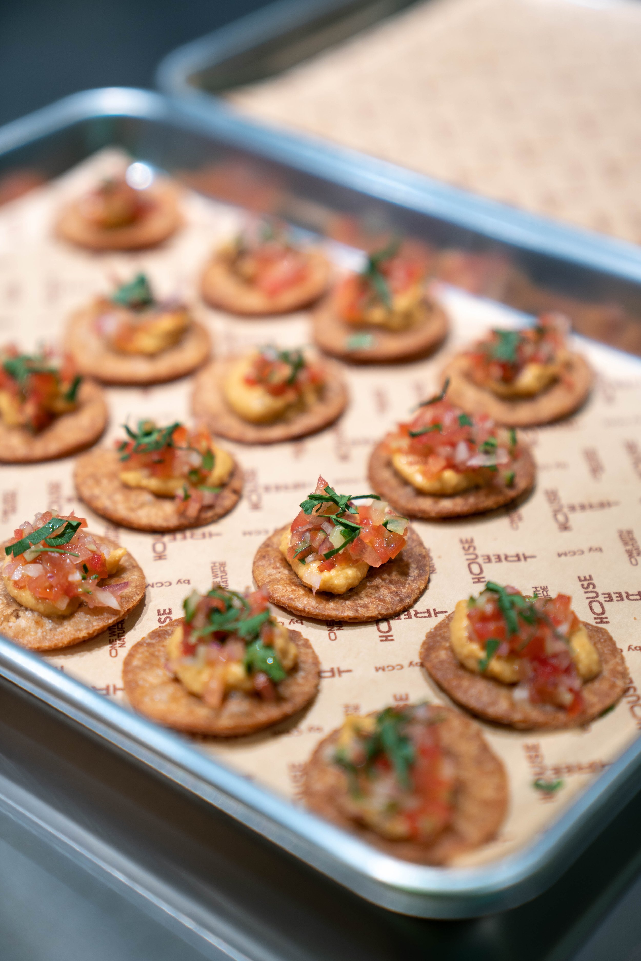 Appetizers on a baking tray featuring round crackers topped with hummus, diced tomatoes, and chopped herbs.