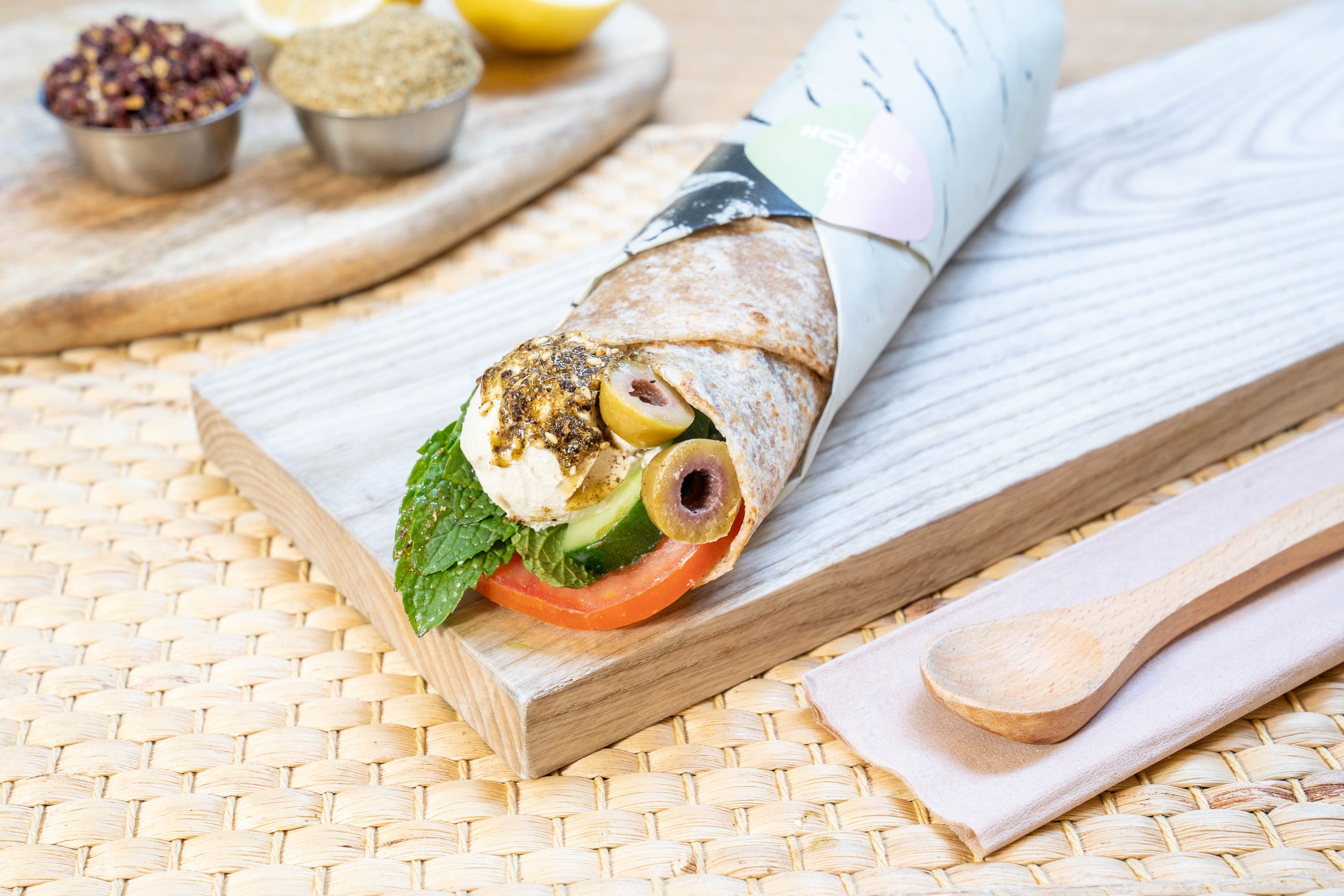 Mediterranean wrap with hummus, olives, cucumber, tomato, and herbs on a wooden board, served with a wooden spoon and spices in the background.