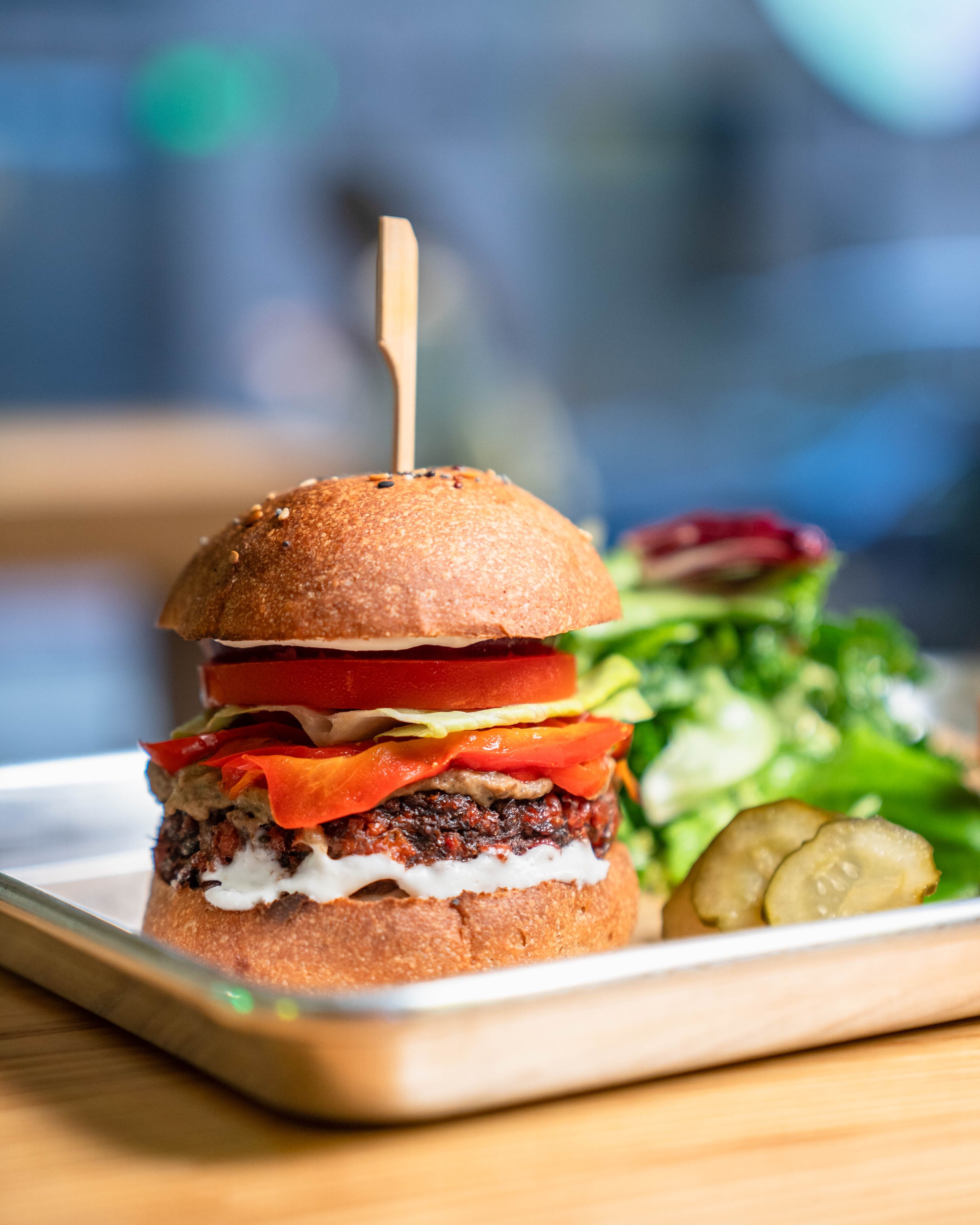 Veggie burger with tomato, lettuce, pepper on a bun, served with greens and pickles on a tray.