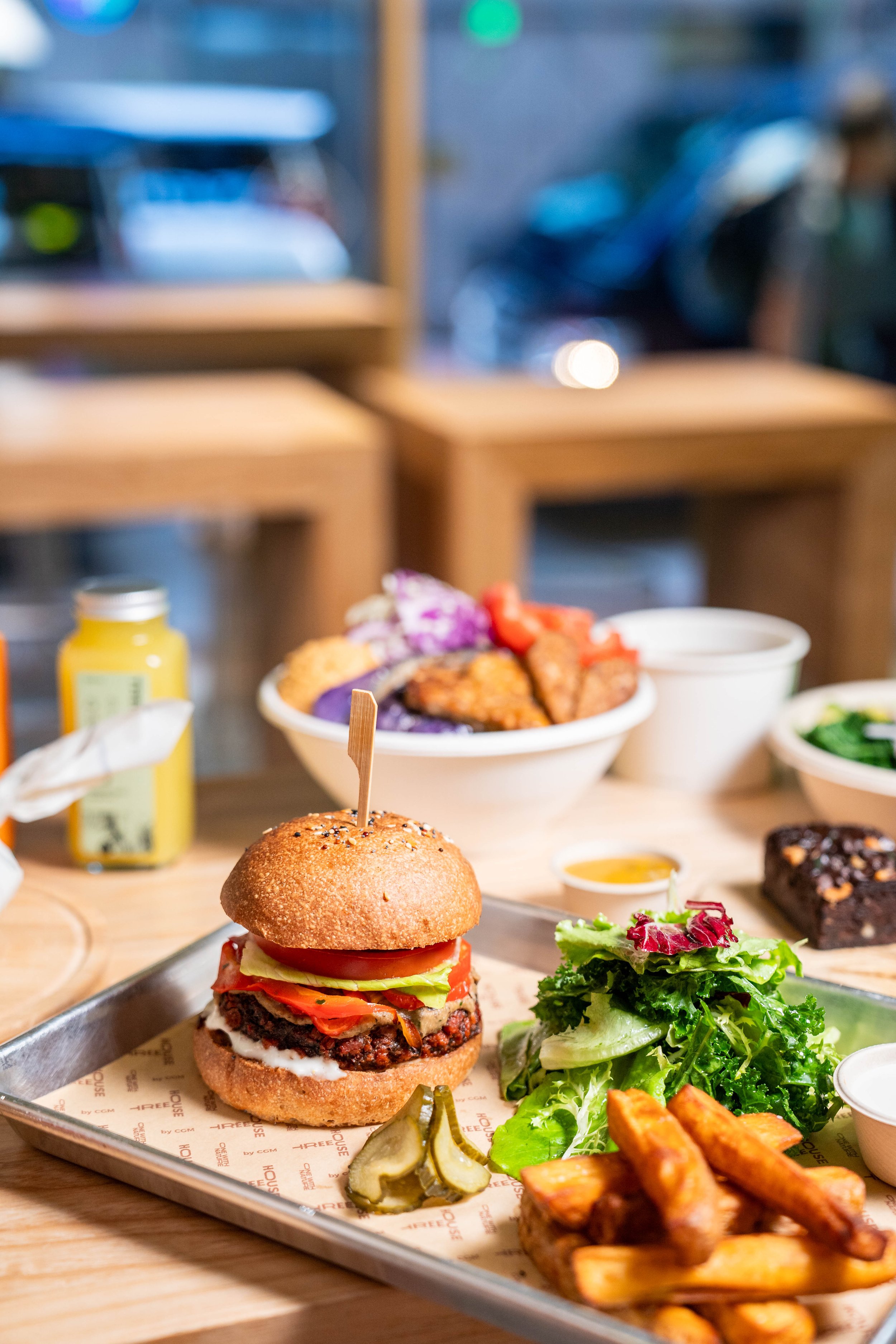 A plant-based burger with toppings on a sesame seed bun, served on a tray with pickles, sweet potato fries, and a side salad. A bottle of juice and a bowl of food are in the background.