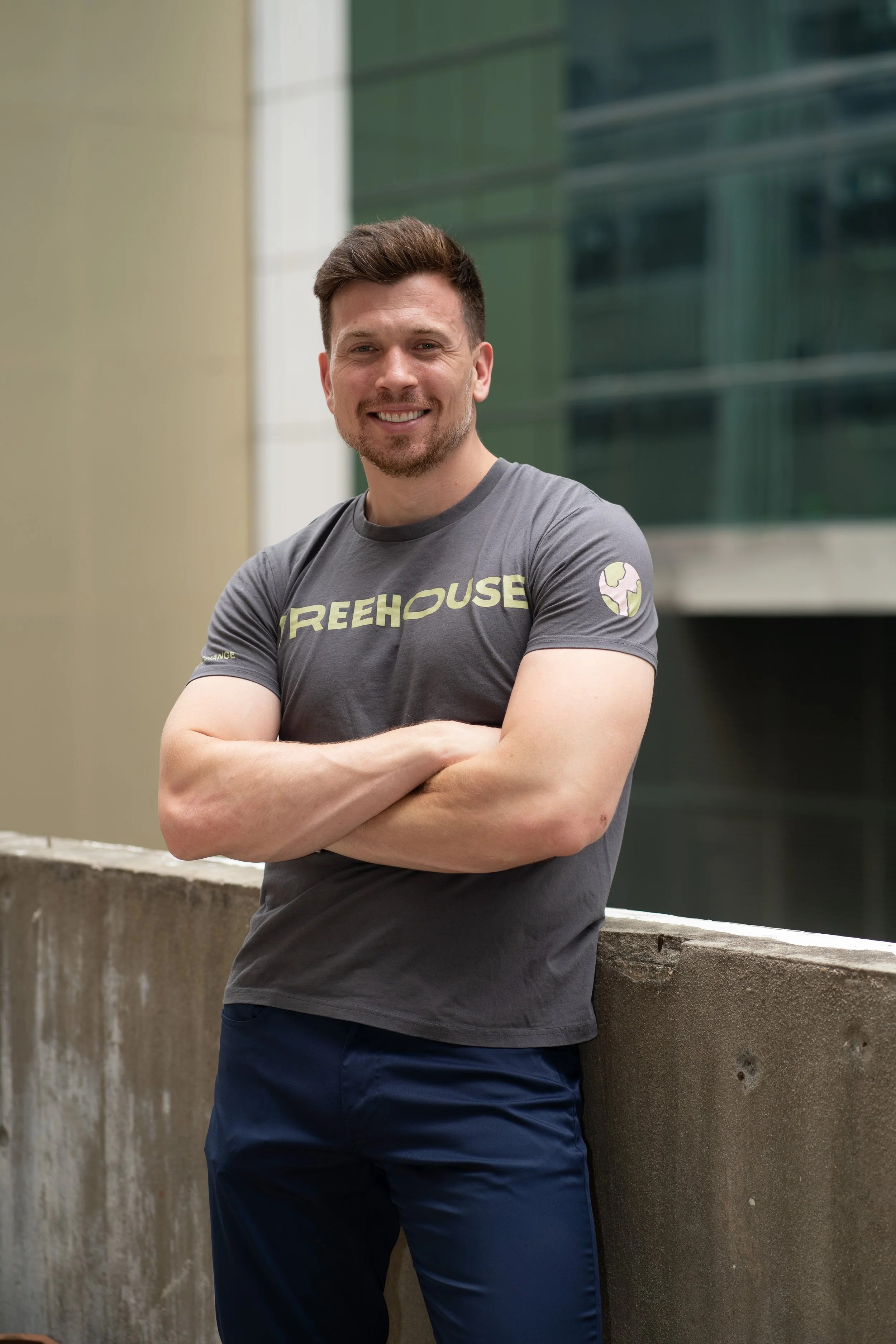 Christian in gray "TREEHOUSE" t-shirt standing with arms crossed outdoors, leaning on concrete railing, with urban background.