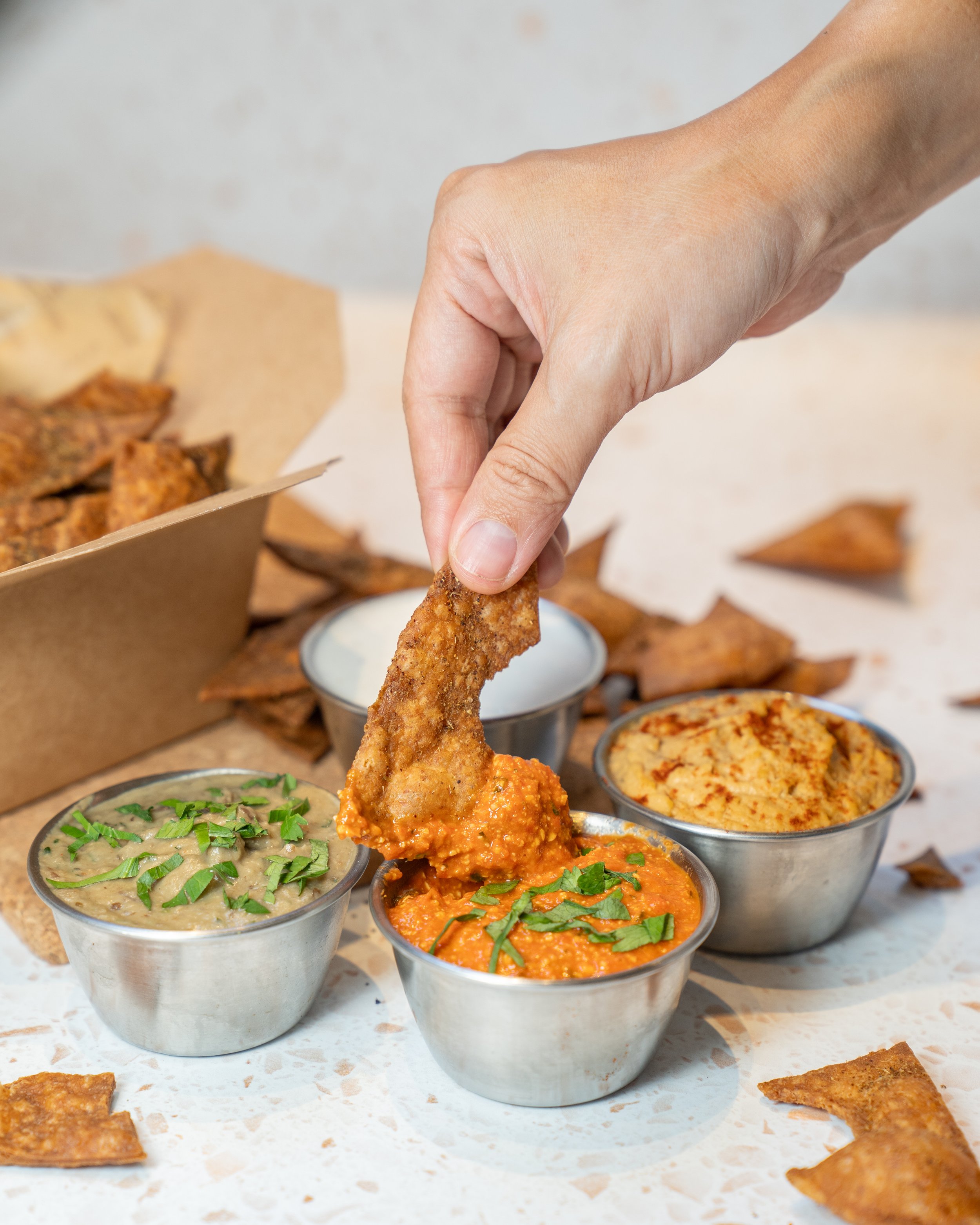 Hand dipping a chip into red dip, with two other dips and chips nearby.